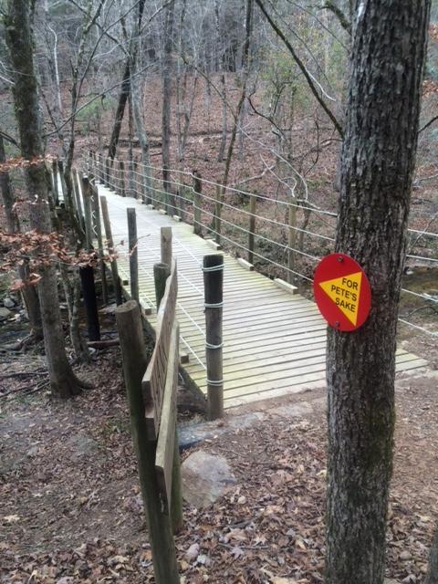 A wooden footbridge spans a shallow creek in a wooded area. The bridge is surrounded by trees with sparse leaves, indicating a late autumn or early winter setting. A red sign reading "FOR PETE'S SAKE" is attached to a nearby tree, while wooden posts and railings line the sides of the bridge, enhancing its rustic charm. Chewacla State Park mountain bike trail.
