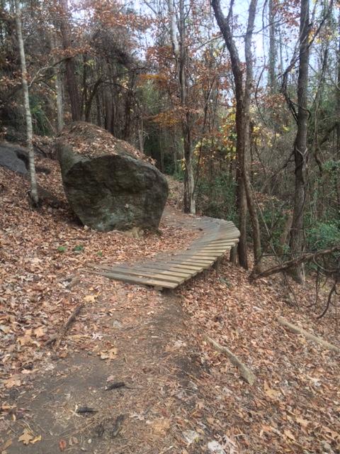 A wooden pathway curving around a large boulder, surrounded by trees with autumn leaves on the ground. The trail is nestled within a wooded area, creating a serene natural setting. Chewacla State Park mountain bike trail.