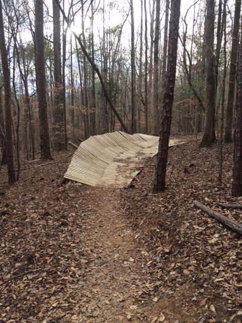 A wooden structure, partially collapsed and lying on the ground, surrounded by trees in a forested area. A dirt trail runs alongside the structure, which is covered in fallen leaves. The scene is set in a quiet, natural environment with bare branches and a cloudy sky above. Chewacla State Park mountain bike trail.