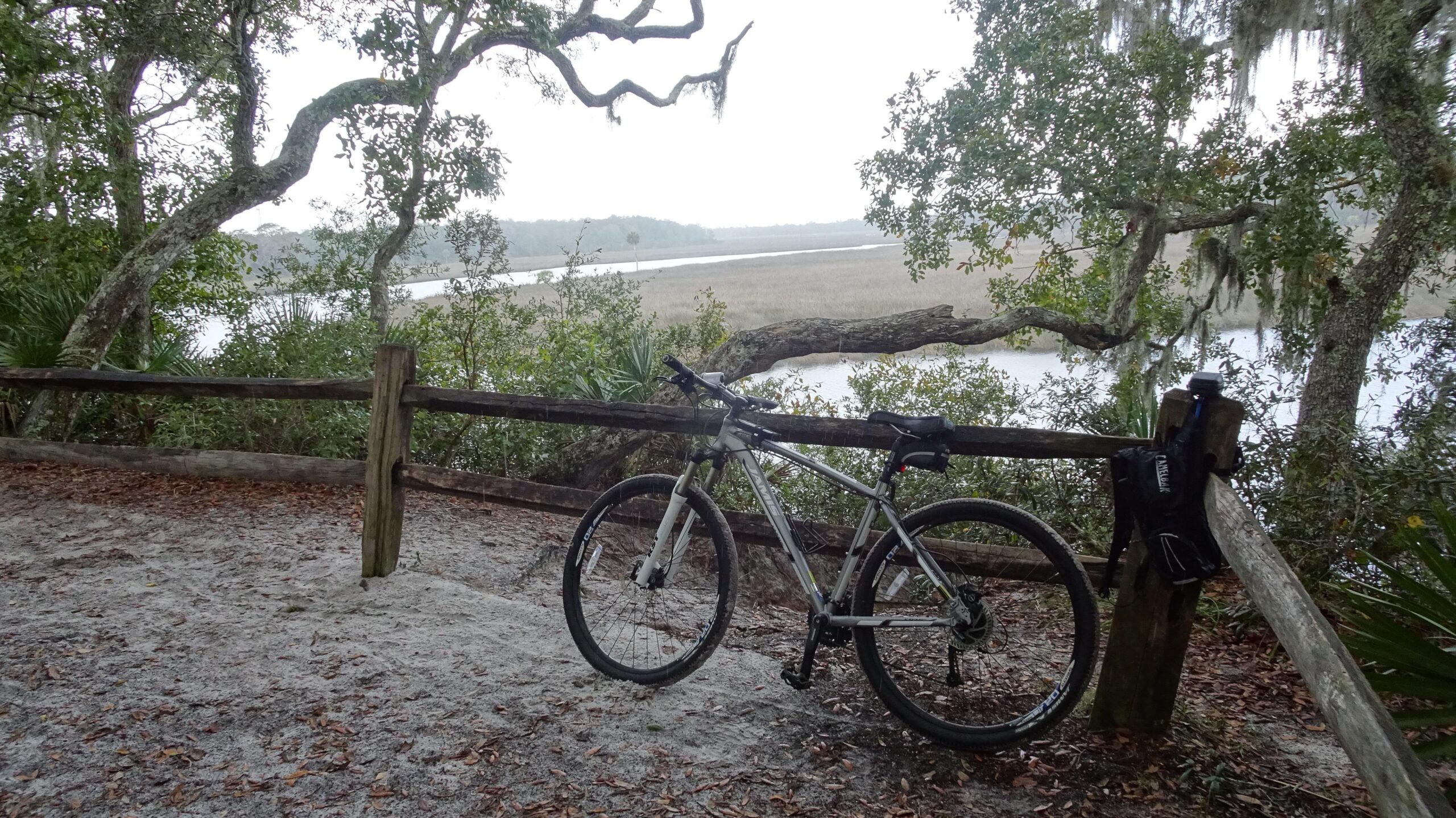 A mountain bike is parked next to a wooden fence overlooking a serene river scene, surrounded by lush greenery and moss-draped trees. The landscape appears misty, suggesting an early morning or overcast day, with tall grasses in the foreground and a calm waterway in the background. Spruce Creek Preserve mountain bike trail.