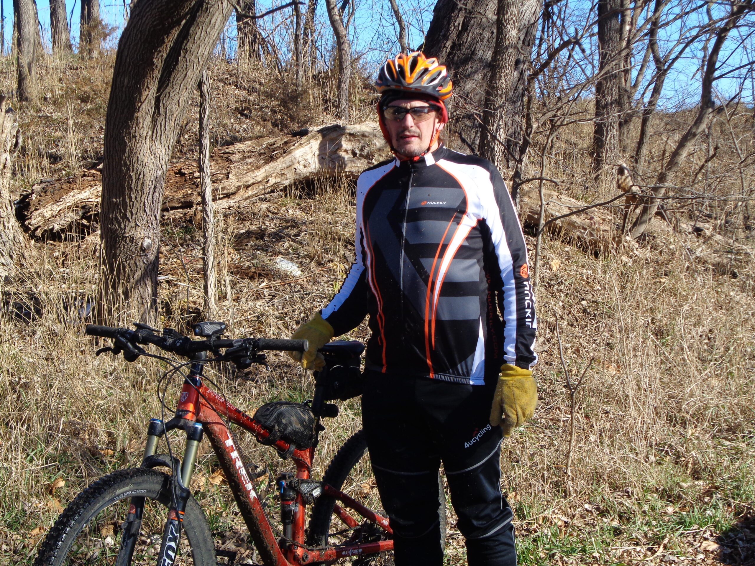 A cyclist wearing a black and orange jersey, sunglasses, and yellow gloves stands next to a mountain bike in a wooded area with dry grass and trees in the background. The scene is set on a clear day with blue skies. Lawrence Riverfront Trails mountain bike trail.