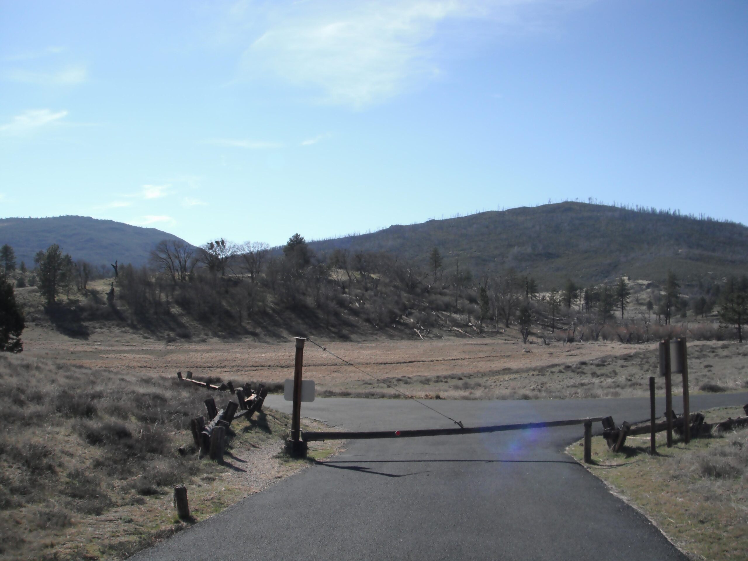 A scenic view of a closed road leading into a natural area, surrounded by hills and sparse vegetation under a clear blue sky. A wooden gate blocks the road, with a backdrop of distant mountains partially covered in trees. The landscape features patches of brown earth and tufts of grass. Anza-Cuyamaca North Side Singletrack mountain bike trail.