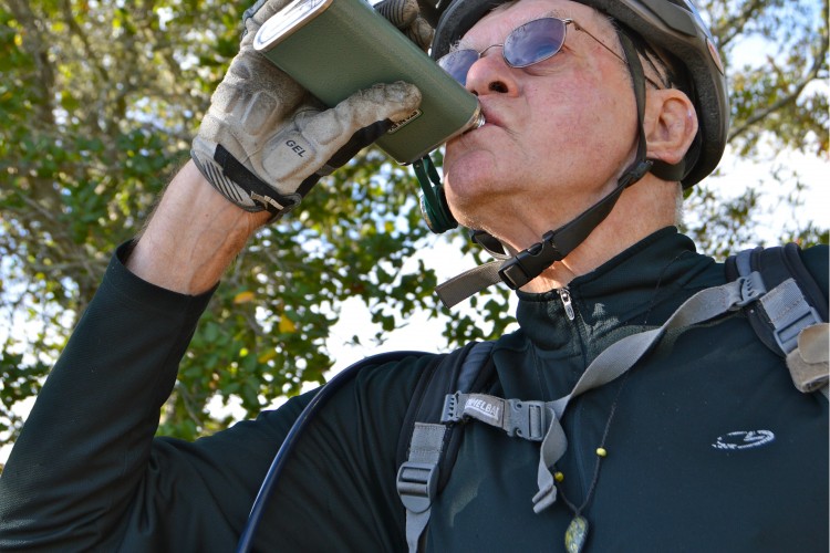 An older man wearing a bicycle helmet and sunglasses drinks from a green container while outdoors. He is dressed in a long-sleeve black shirt and has a backpack with a hydration tube visible. Surrounding him are trees with green foliage, indicating a natural setting.