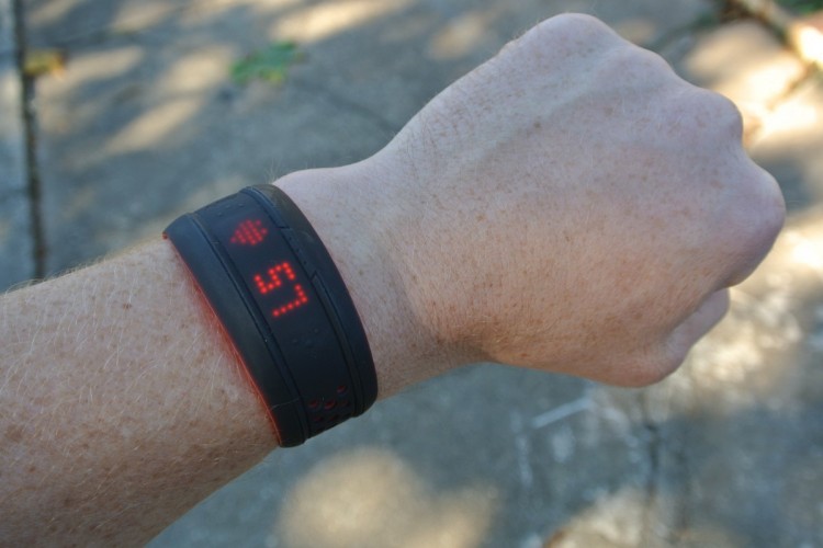 A close-up of a wrist wearing a black fitness tracker displaying a heart rate of 57. The wrist is positioned against a blurred outdoor background, with sunlight creating shadows on the surface.