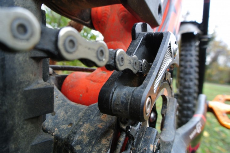 Close-up view of a mountain bike's drivetrain, featuring the chain, rear derailleur, and components coated in dirt, set against a blurred green background.
