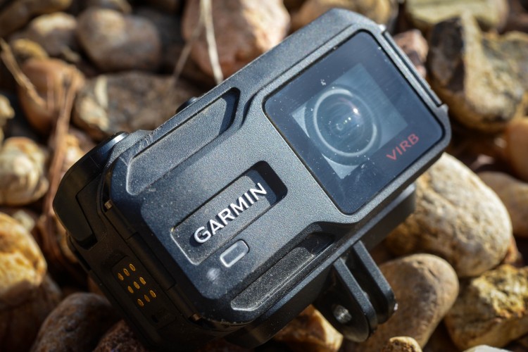 A Garmin VIRB action camera resting on a bed of small, smooth pebbles, featuring a sleek black design with a lens and control buttons visible.
