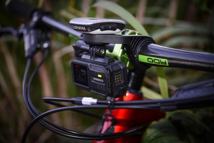 Close-up of a bike's handlebars featuring a mounted action camera and GPS device, surrounded by green foliage. The bicycle has a red frame with black and green components.