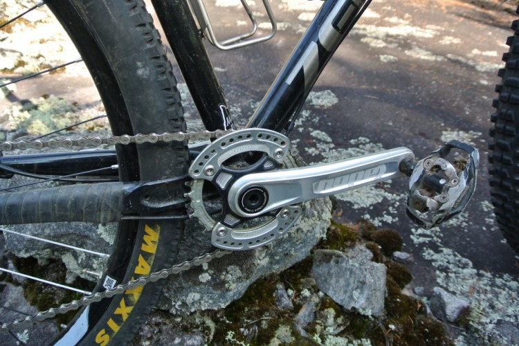 Close-up view of a mountain bike's drivetrain, featuring the chain, crankset, and pedals, with a rugged terrain background. The bike's tire is partially visible, along with mossy rocks on the ground.
