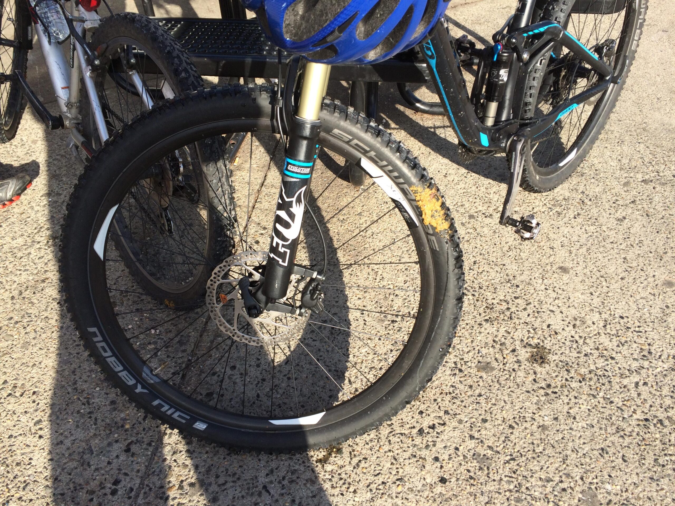 A close-up image of a mountain bike tire and front fork, showing a partially visible disc brake and other bikes in the background. The tire features a knobby tread design, and there is mud or dirt on the sidewall. The surface below is concrete. Trails seperated by streets mountain bike trail.