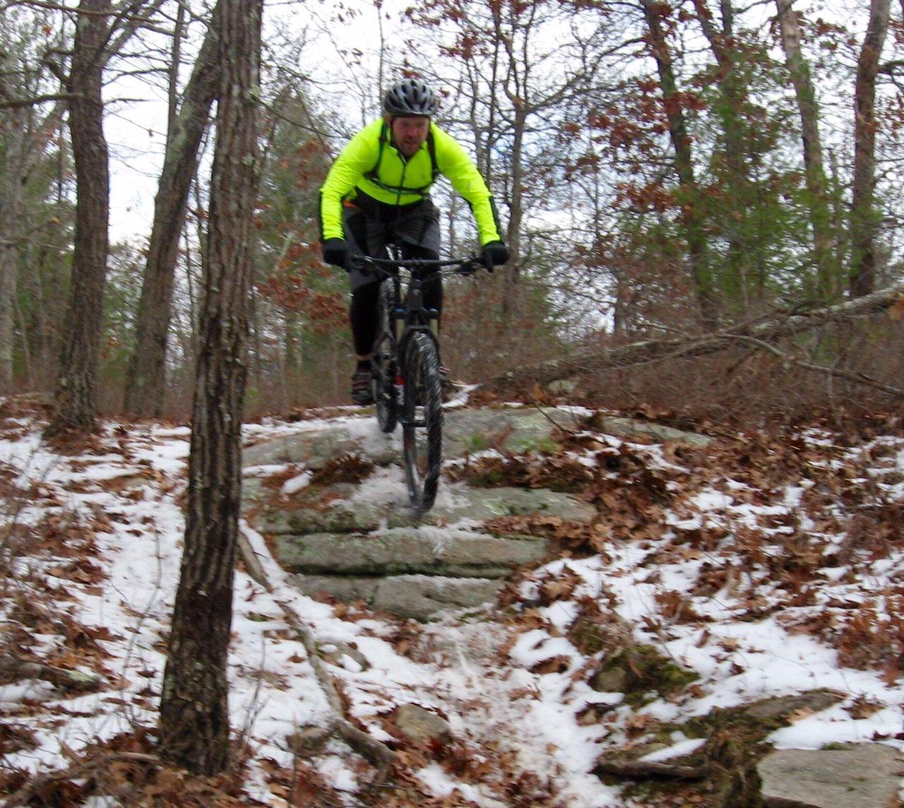 A mountain biker in a bright yellow jacket navigates over rocky terrain during winter, surrounded by trees and fallen leaves, with patches of snow on the ground. Douglas State Forest mountain bike trail.