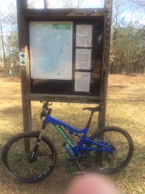 A mountain bike leaning against a wooden information board displaying a map and details about a park, surrounded by grassy terrain and trees in the background. Flat Rock Park mountain bike trail.