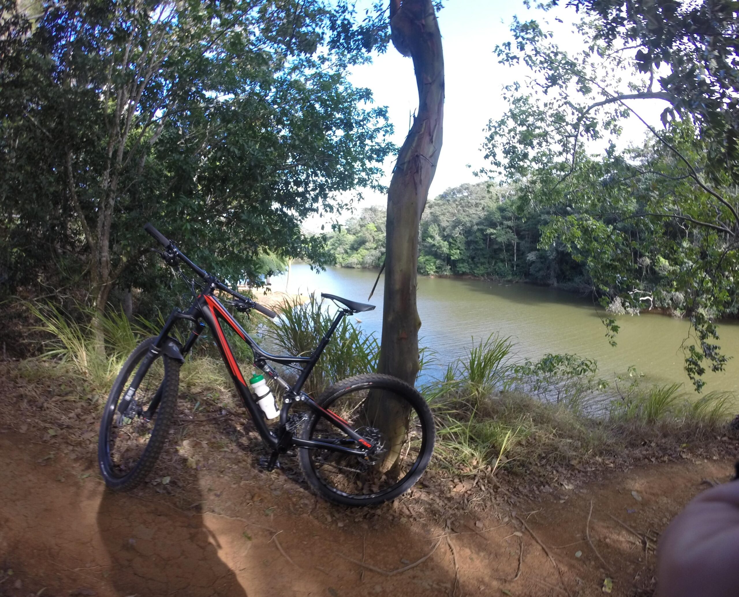 Mountain bike resting near a riverbank surrounded by lush greenery and trees under a clear blue sky. Hacienda Sabanera mountain bike trail.