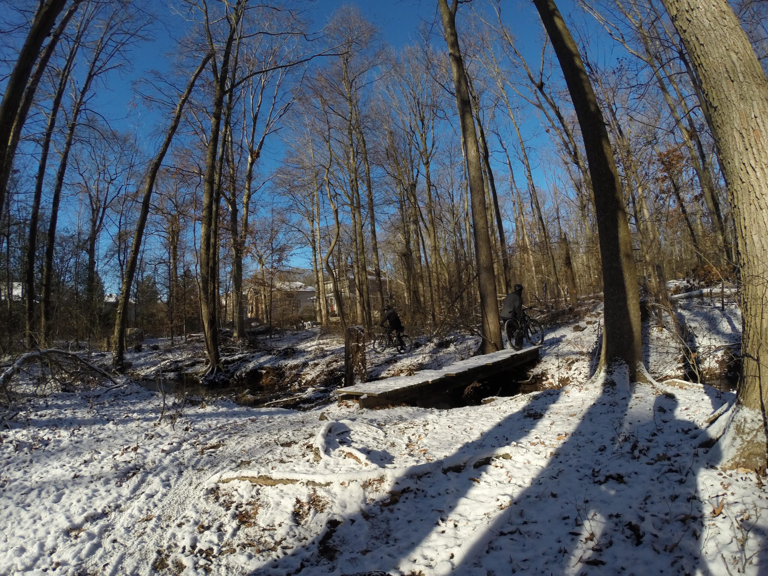 A snowy forest scene with a clear blue sky, showing bare trees and a small creek. Two mountain bikers are seen on a wooden bridge crossing over the creek, while shadows of trees and cyclists stretch across the snow-covered ground. Trails seperated by streets mountain bike trail.