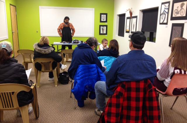 A small group of people is seated in a classroom setting, facing a speaker standing at a table equipped with materials for a presentation. The wall behind features artwork, and the room has a vibrant green wall section. Attendees are wearing jackets, indicating a chilly environment, and some are holding papers, likely related to the topic being discussed. The atmosphere appears focused and engaged.
