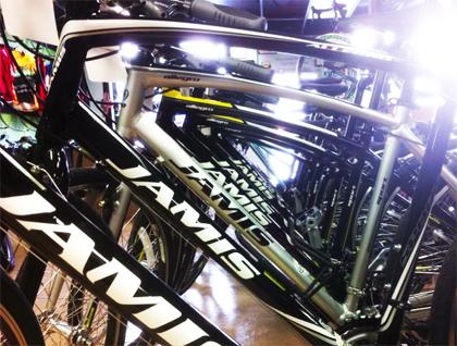 Close-up view of multiple Jamis bicycles lined up in a store, showcasing their frames and branding. The bikes have a mix of black and silver colors, with reflections and highlights illuminating the scene.