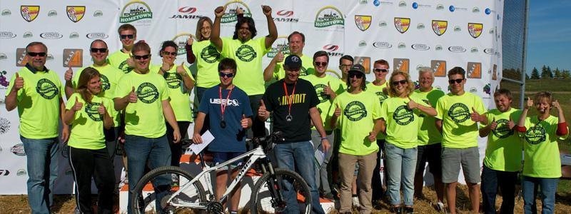A group of enthusiastic people wearing bright yellow t-shirts stands together, smiling and giving thumbs up in front of a promotional backdrop. A mountain bike is displayed in the foreground, and the atmosphere suggests a celebration or award ceremony related to cycling.
