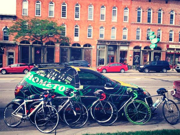 A lineup of five road bicycles parked next to a colorful car featuring the words "HOMETOWN" and design elements. The setting includes a brick building with multiple windows in the background, and a few parked cars in the street. Trees are visible along the sidewalk, creating a vibrant urban scene.