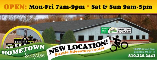 Image of a sign for Hometown Bicycles, featuring the text "OPEN: Mon-Fri 7am-9pm, Sat & Sun 9am-5pm" and "NEW LOCATION! Bicycle Adventure Center." The sign includes the address: 10595 Grand River, Brighton, MI 48116, and a contact number: 810.225.2441. The background shows a building with trees and an outdoor setting.