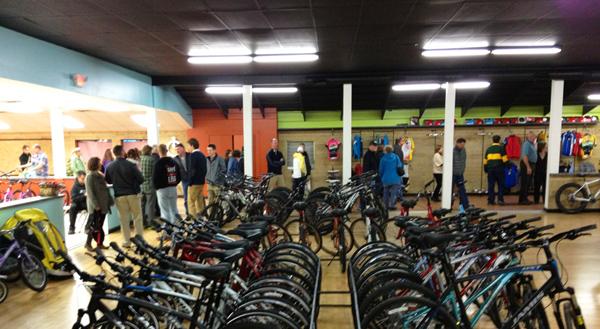 A busy indoor bicycle shop featuring a row of bicycles lined up in the foreground. In the background, a group of people are gathered at a counter, while others browse clothing and accessories displayed on the walls. The shop has brightly colored walls and a well-lit atmosphere.