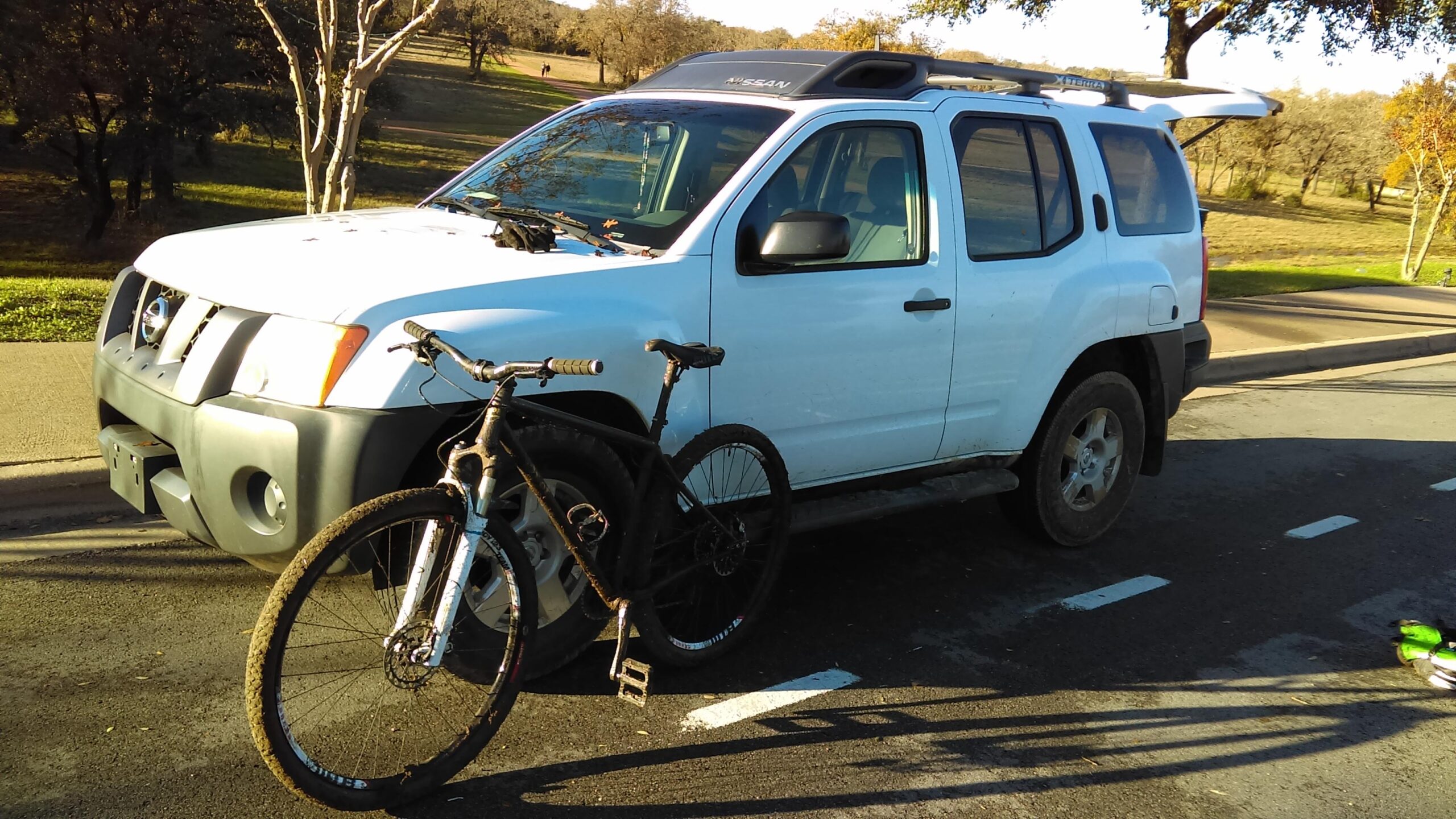Spot Honey Badger Geared: A white Nissan SUV parked on a road with a mountain bike resting against the front. The bike has a black frame and appears to be muddy, suggesting recent use. In the background, a grassy area with trees is visible, indicating an outdoor setting.