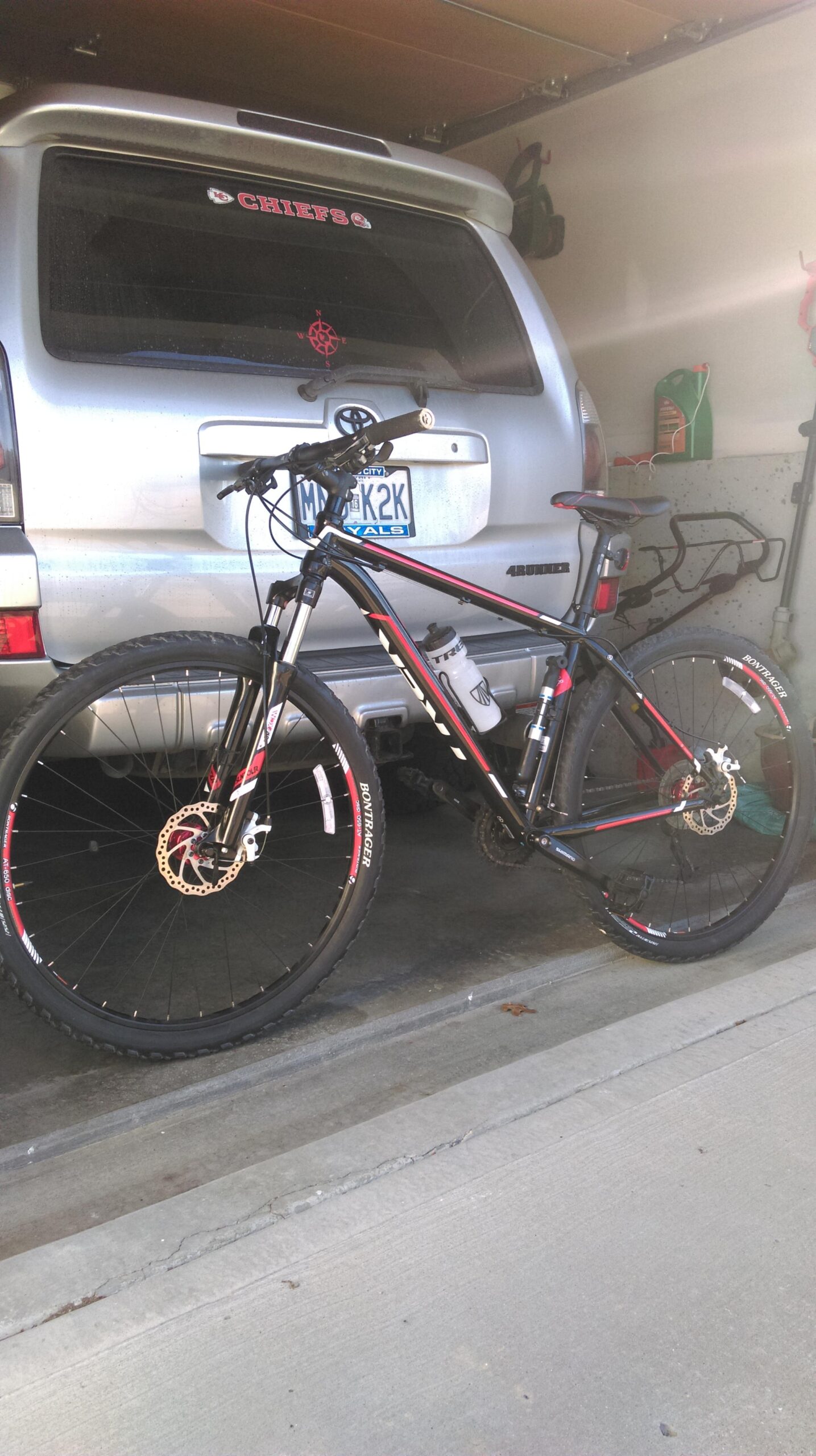 Trek Marlin: A mountain bike parked in a garage next to a silver SUV. The bike features black and red colors, with disc brakes and a water bottle holder attached. The SUV has a "Chiefs" sticker on the rear window, and various items are visible on the garage wall.