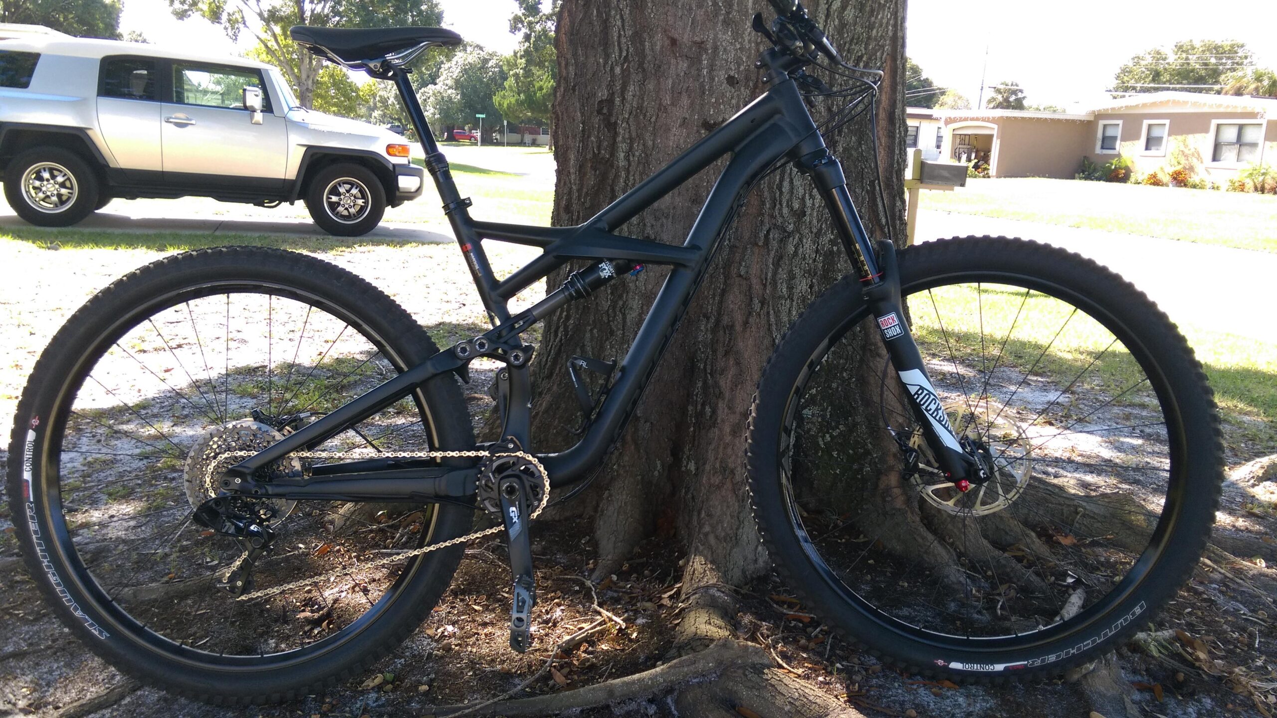 Specialized Enduro Comp 29: A black mountain bike leaning against a tree, with a silver SUV parked in the background. The bike features wide tires, a suspension system, and a chain drivetrain. The setting is a sunny outdoor area with grass and a paved road.