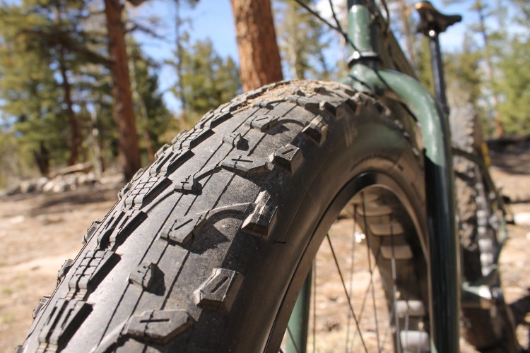 Close-up view of a mountain bike tire with an intricate tread pattern, showcasing its rugged design. In the background, trees and a natural landscape are visible, indicating an outdoor setting.