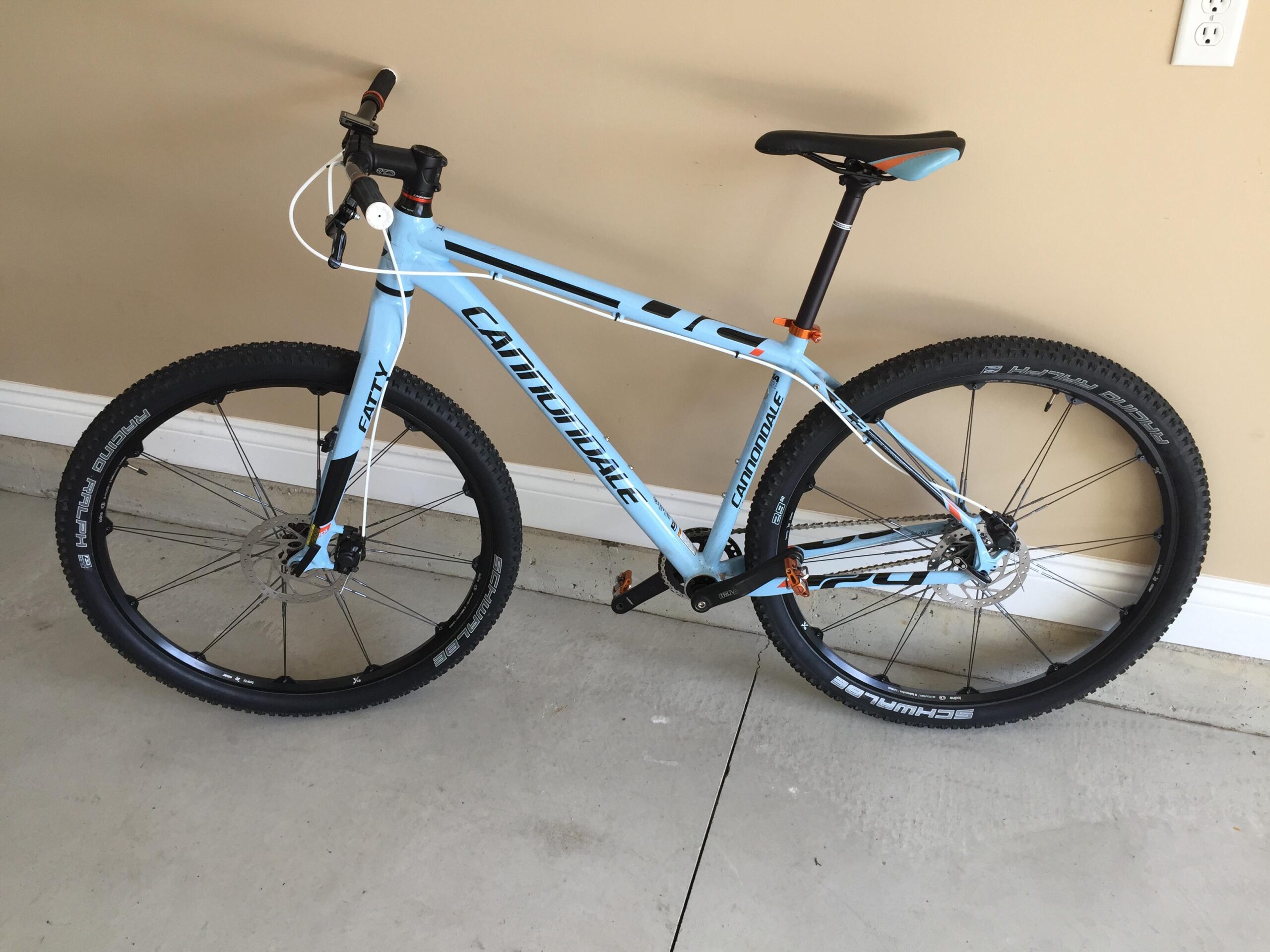 Cannondale Trail SL 29 SS: A blue Cannondale mountain bike with wide tires, featuring a black seat and handlebars, positioned on a concrete surface against a beige wall. The bike has orange accents and is equipped with disc brakes.