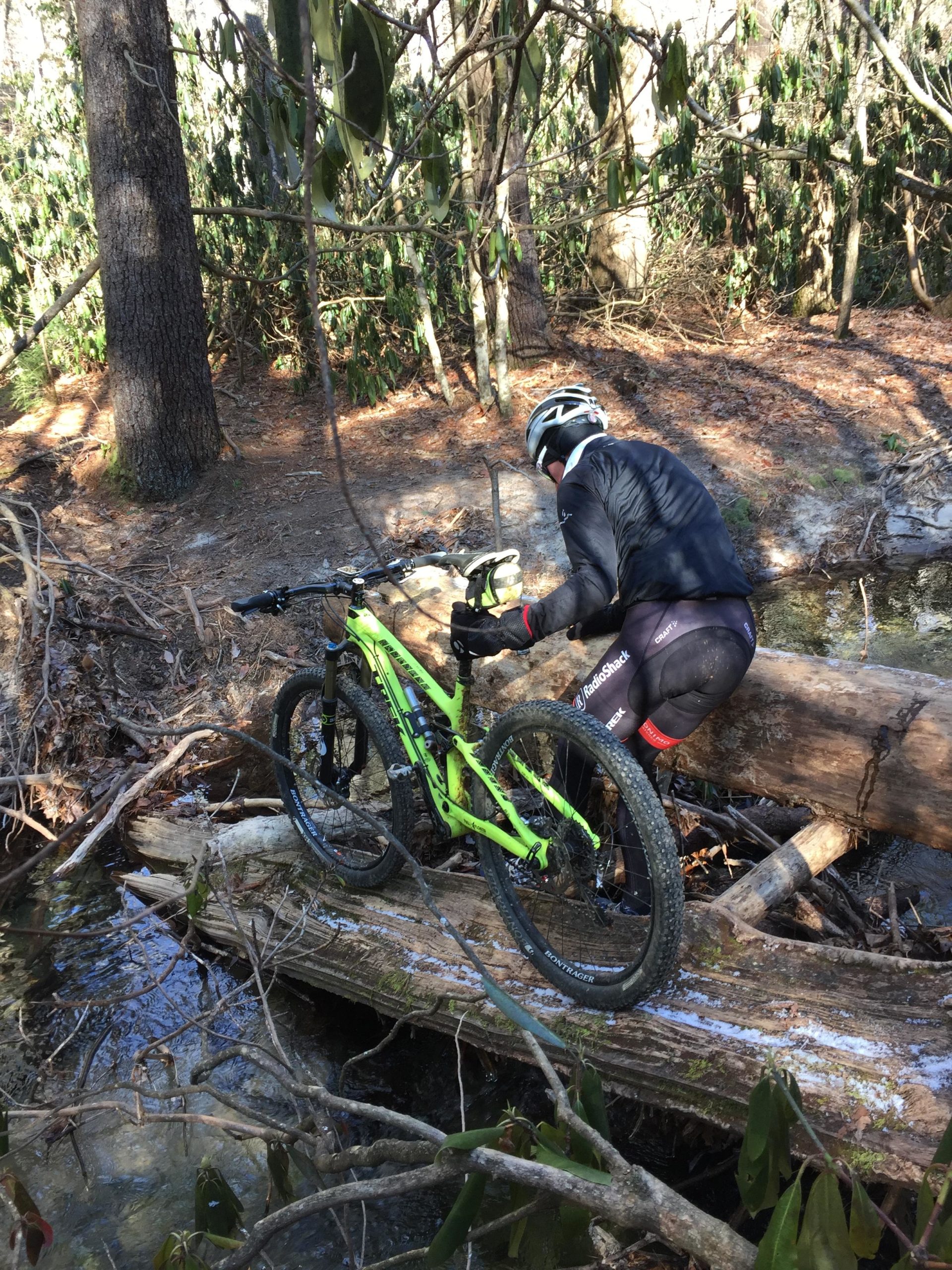 A cyclist navigating across a log bridge over a small stream in a wooded area. The cyclist is wearing a helmet and cycling gear, and their bright green mountain bike is positioned on the log. Surrounding trees and foliage create a natural, rugged setting. DuPont State Forest mountain bike trail.