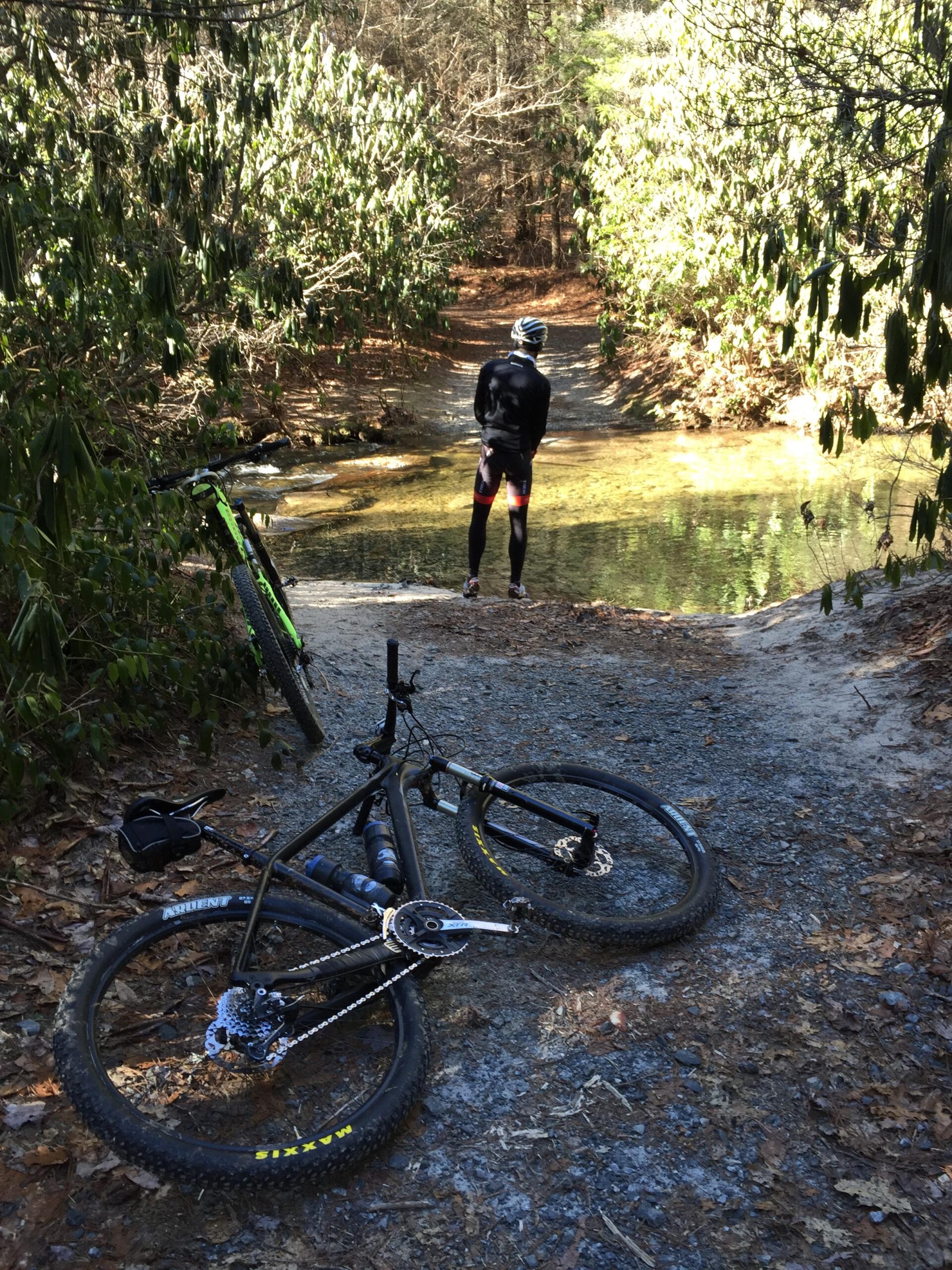 Foundry Cycles Tomahawk: A mountain biker stands near a clear stream surrounded by lush greenery. Two bikes are positioned on the gravel path in the foreground, with one bike lying on its side. The scene captures a peaceful moment in nature, showcasing the beauty of outdoor cycling.