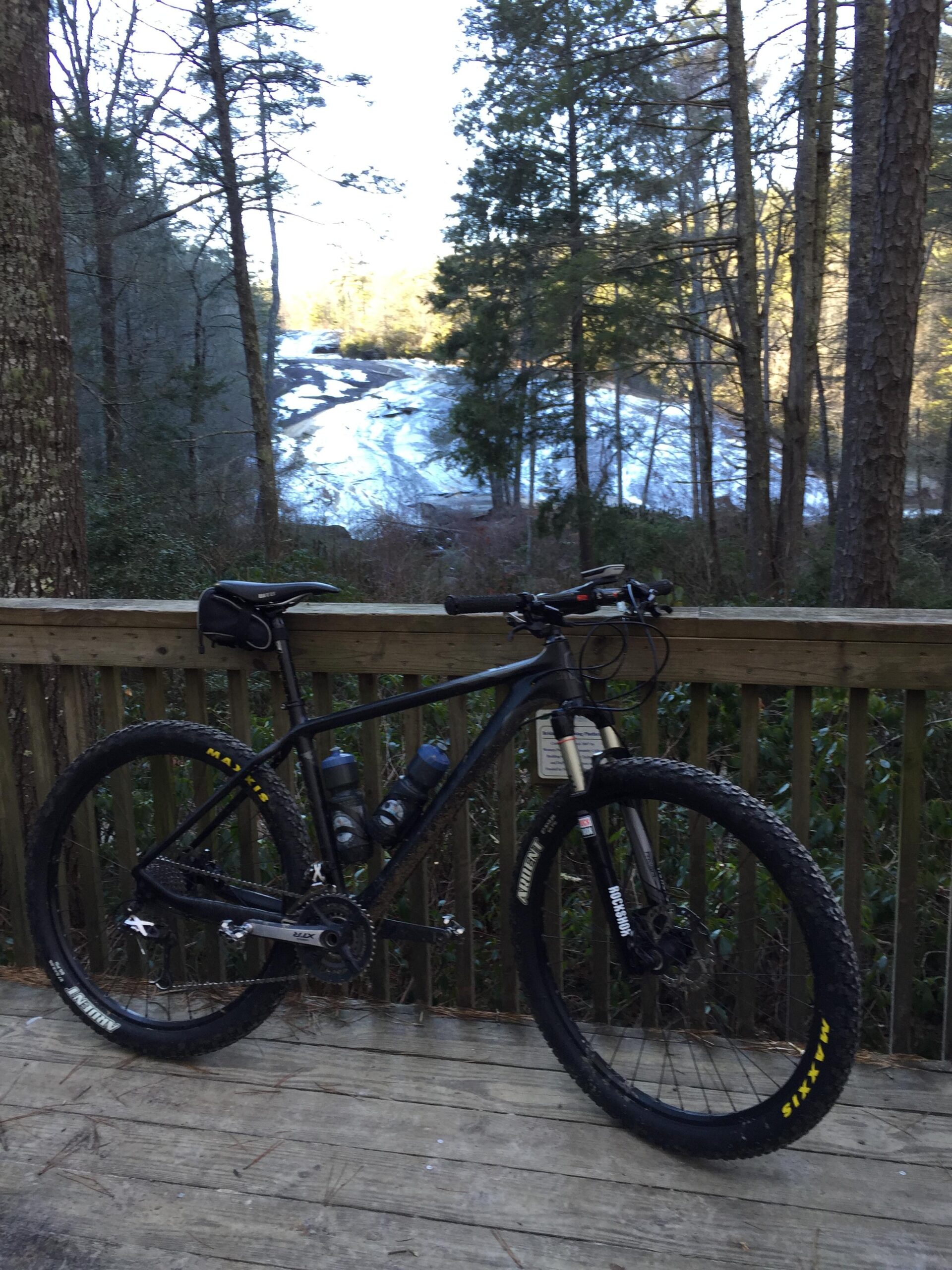 Foundry Cycles Tomahawk: A black mountain bike is parked on a wooden deck, with two water bottles attached to its frame. In the background, a scenic view of a rocky landscape is visible, flanked by tall trees and a gentle stream cascading down the rocks. The setting suggests a tranquil, outdoor environment, ideal for cycling and enjoying nature.