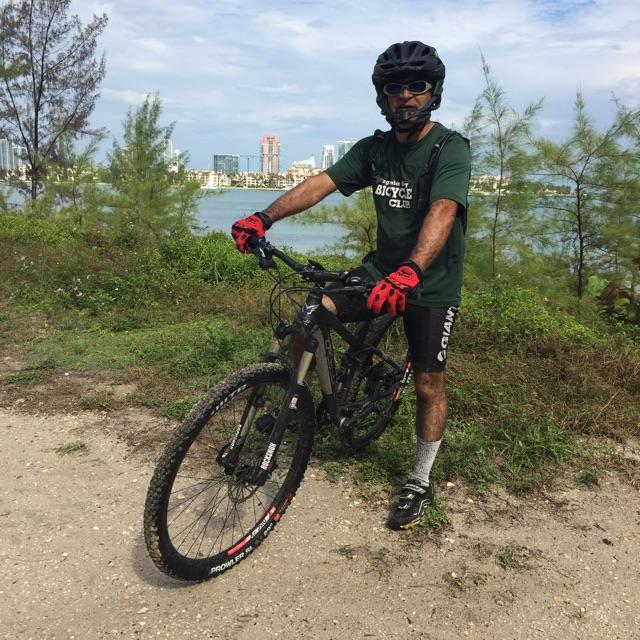 A person in a green Bicycle Club t-shirt and black shorts is standing next to a mountain bike on a dirt path. They are wearing a black helmet and red gloves, with a scenic view of a body of water and city skyline in the background. The surroundings include green vegetation and trees under a partly cloudy sky. Virginia Key North Point mountain bike trail.