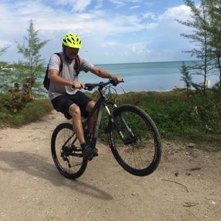 A person wearing a bright yellow helmet performs a wheelie on a mountain bike along a sandy path by the water, with trees and a blue sky in the background. Virginia Key North Point mountain bike trail.