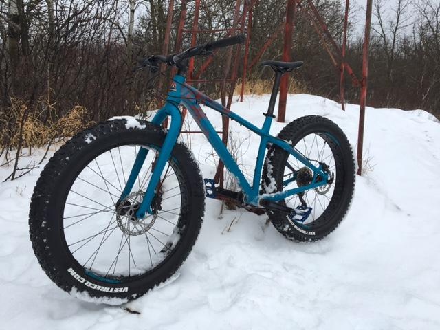 Rocky Mountain Blizzard: A blue fat bike with large tires standing on snowy terrain, surrounded by sparse vegetation and a metal structure in the background. The bike's frame is angled to show its distinctive design and wide tires suitable for winter riding.