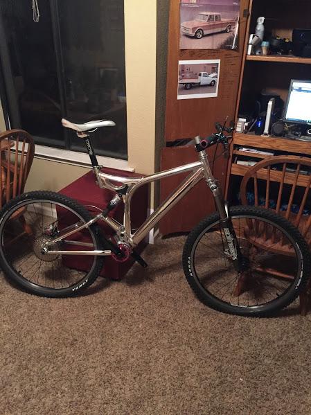 GT xcr le: A silver mountain bike is positioned in a room with wooden chairs and a desk in the background. The bike features thick tires and a prominent front suspension fork. A window nearby allows natural light to enter, and there is a small piece of furniture and a wall with attached pictures visible in the scene. The room has a cozy, lived-in feel.