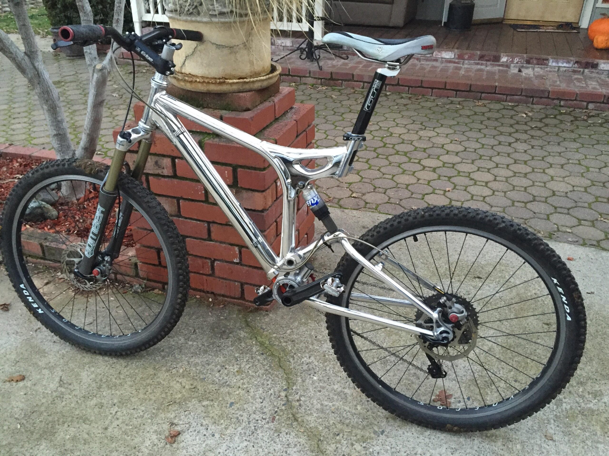 GT xcr le: A silver mountain bike parked beside a brick planter, featuring a suspension fork, knobby tires, and a sleek frame design. The background shows a stone walkway and a glimpse of a porch area.