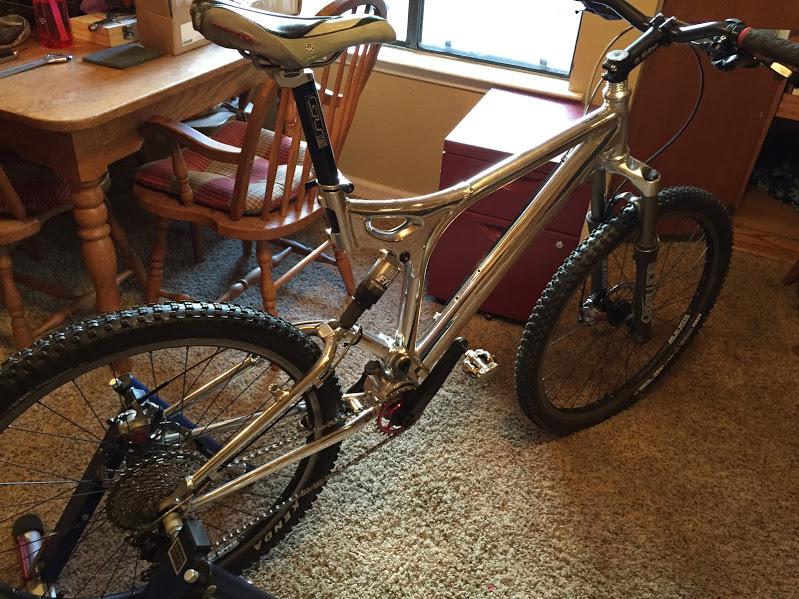 GT xcr le: A silver mountain bike is positioned indoors, showcasing its sleek frame and sturdy tires. The bike has a visible rear suspension, a comfortable seat, and various components including gears and pedals. In the background, there's a wooden dining table with chairs and a red storage box, indicating a cozy room setting.