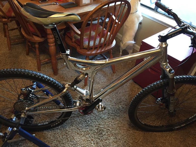 GT xcr le: A silver mountain bike with a sleek frame, featuring knobby tires and a visible suspension system. The bike is positioned indoors on a carpeted floor, with wooden dining chairs and a red storage box in the background.