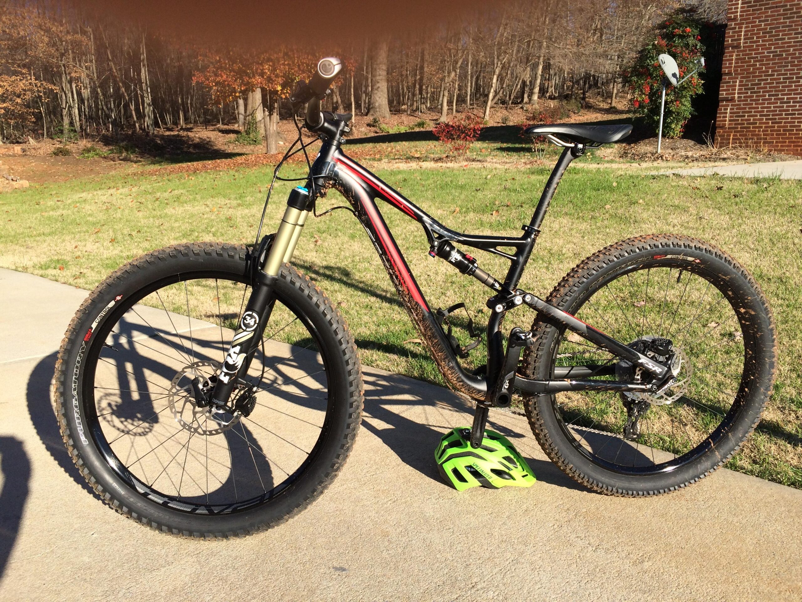 Specialized Stumpjumper FSR 6Fattie: A mountain bike with a black and red frame, equipped with wide tires and a suspension fork, is parked on a concrete surface. The bike features a green pedal and is surrounded by a grassy area with trees in the background, suggesting an outdoor setting.