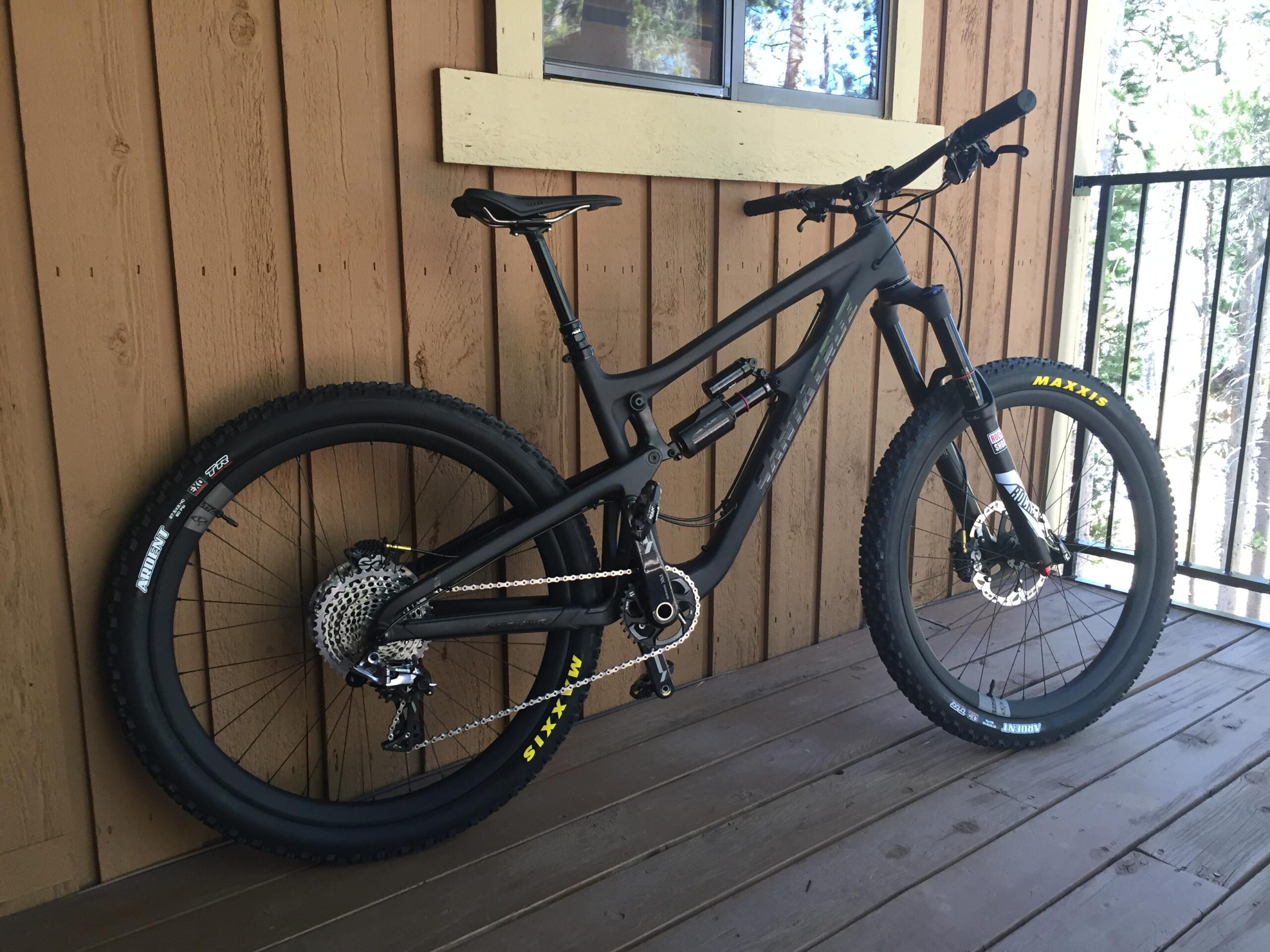 Santa Cruz Nomad: A black mountain bike leaning against a wooden wall. The bike features thick tires with "Maxxis" branding, a prominent suspension system, and a sleek design suitable for off-road riding. The background includes a portion of a balcony railing and a window, suggesting a forested or outdoor setting.
