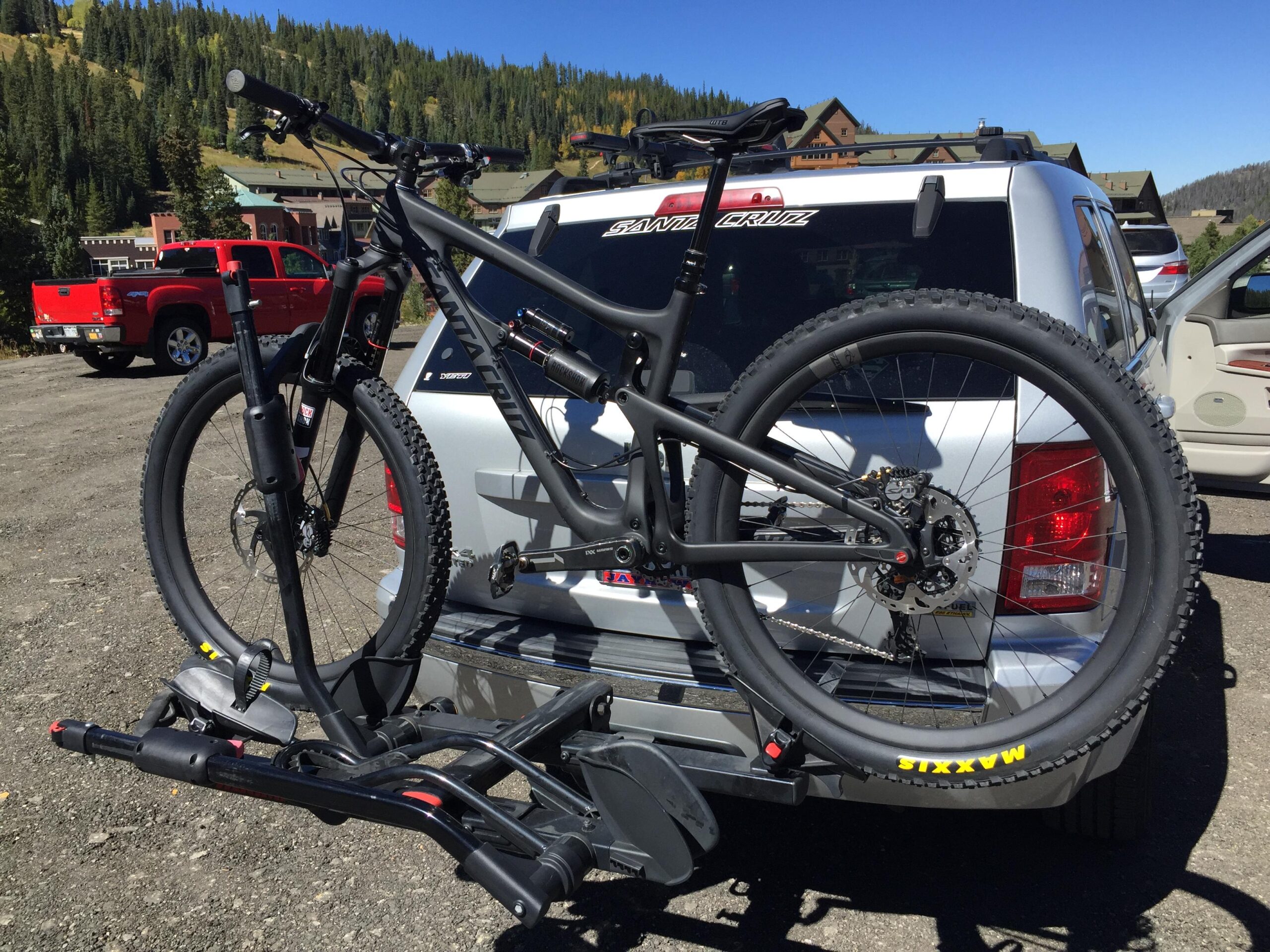 Santa Cruz Nomad: A black mountain bike is secured on a bike rack attached to the back of a silver SUV, parked in a gravel area. The backdrop features a mountainous landscape with trees and distant buildings, under a clear blue sky. The bike is positioned upright, showcasing its tires and frame.