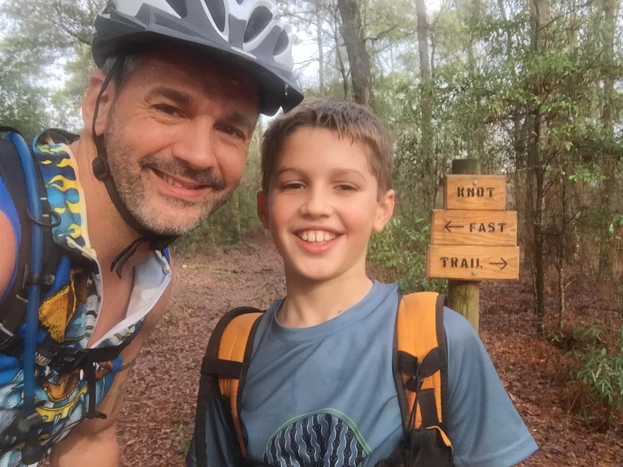 A smiling man and boy pose for a selfie on a forested mountain biking trail. The man wears a helmet and a sleeveless shirt, while the boy is wearing a t-shirt and an orange backpack. Behind them, a wooden sign indicates directions for "Knot" and "Fast" trails. The Knot mountain bike trail.