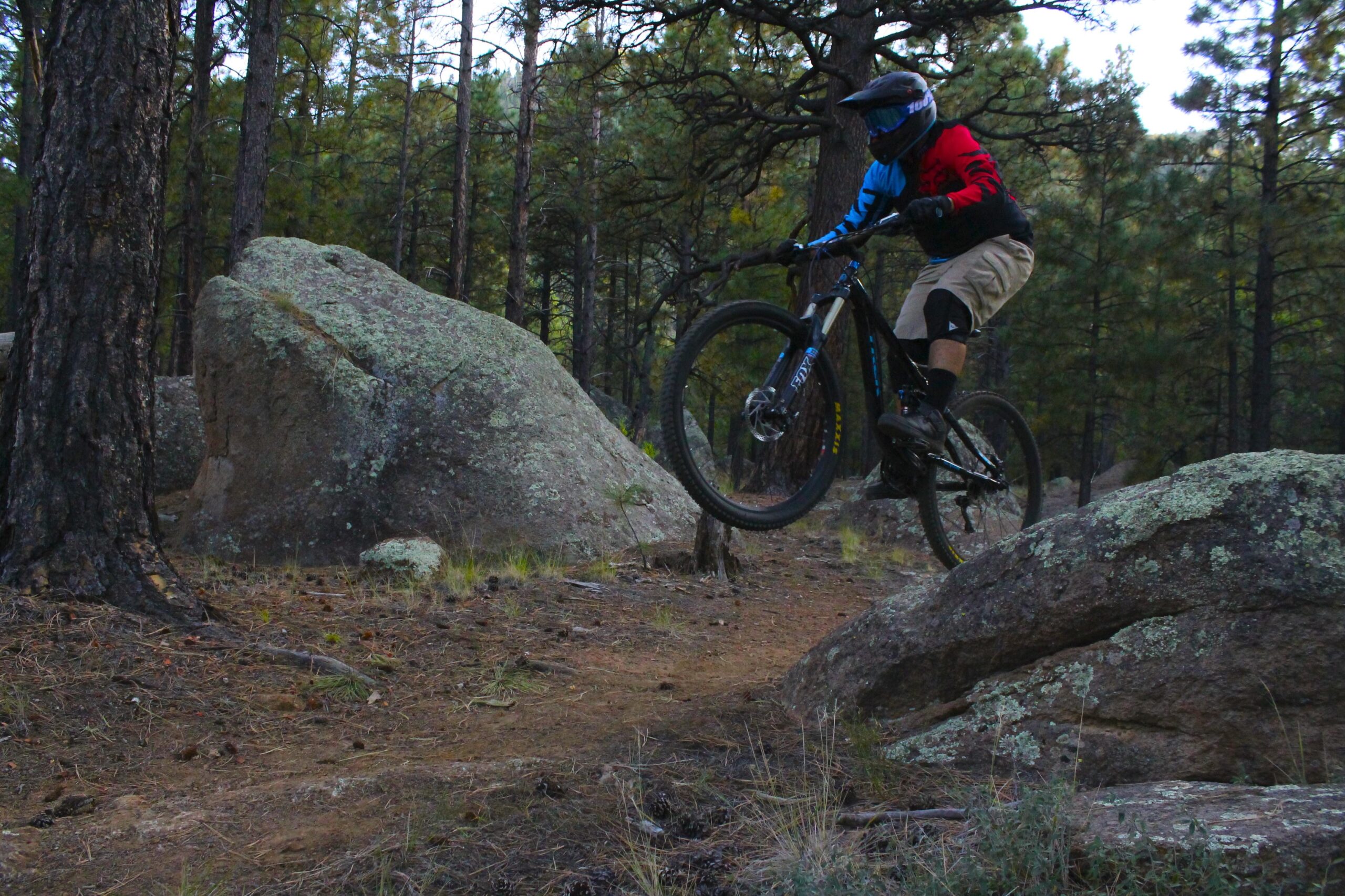 Trek Remedy 8 29: A mountain biker performing a jump over a large rock on a forest trail, surrounded by tall trees and rocky terrain. The rider is wearing a helmet and protective gear, showcasing an athletic posture in mid-air.