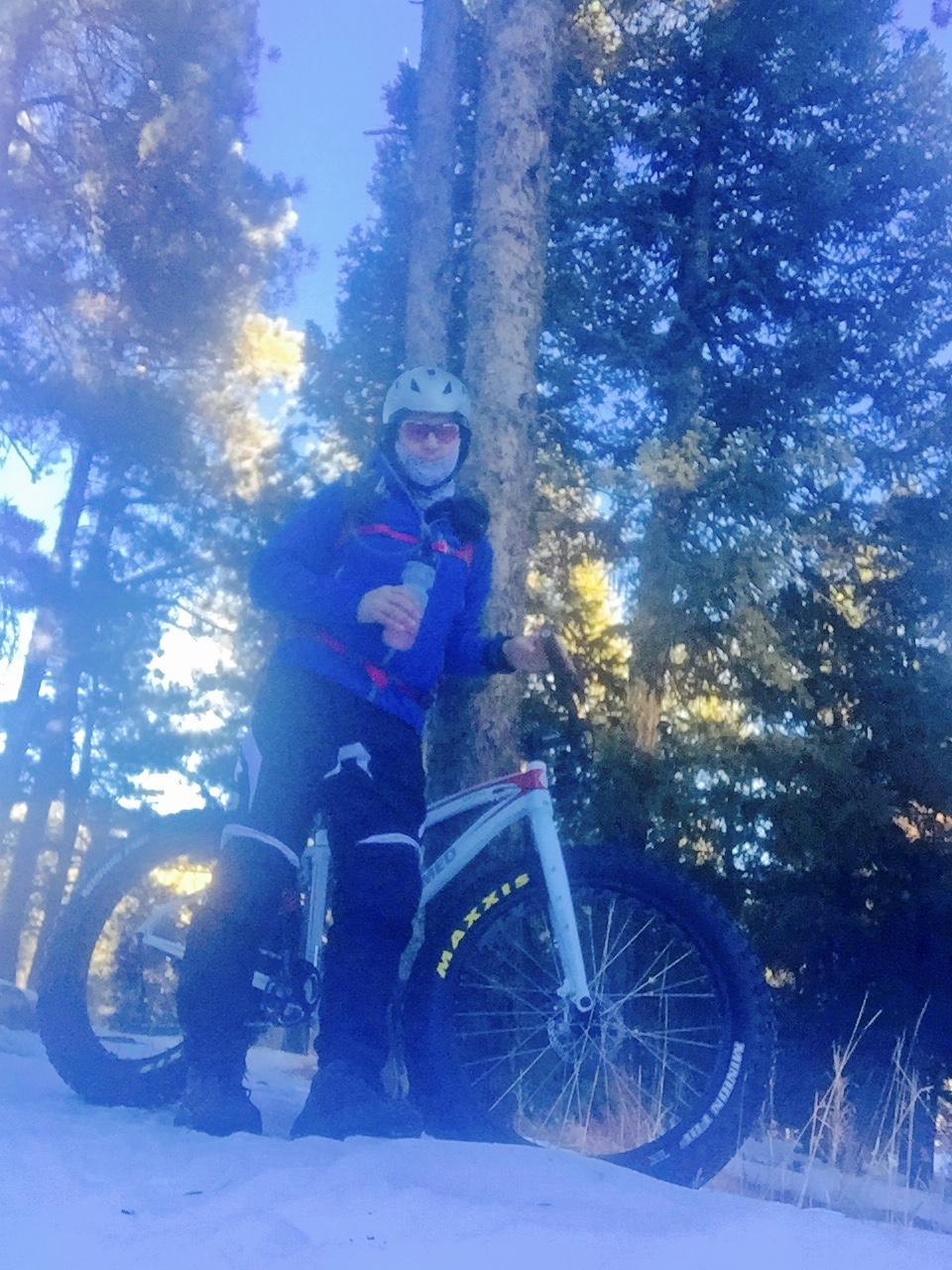 Framed Framed Wolftrax: A person wearing a helmet and sunglasses stands next to a white fat bike in a snow-covered forest. They are dressed in a blue jacket and black pants, holding a drink in one hand, surrounded by tall trees and snow. The scene captures a winter outdoor activity.