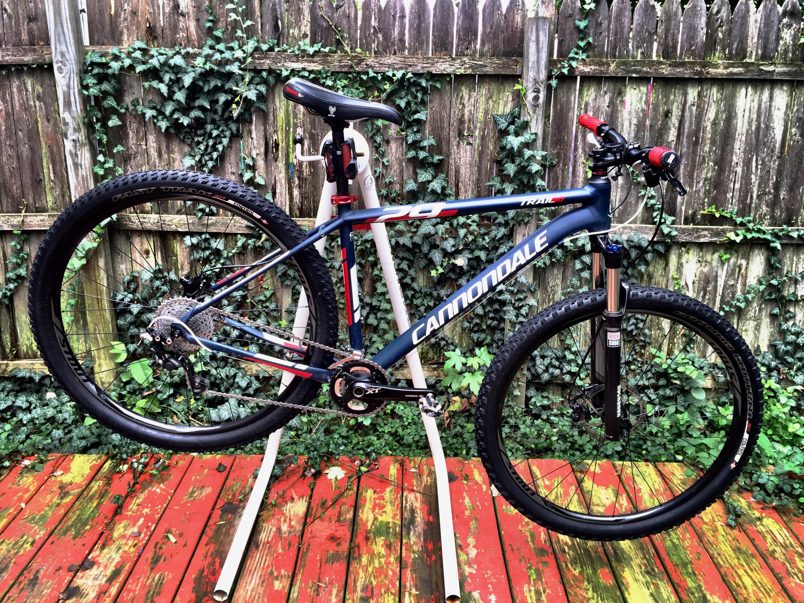 Cannondale Trail 5: A Cannondale mountain bike is displayed on a bike stand, showcasing its sleek design and components. The bike features a blue and red frame, black wheels, and is surrounded by a wooden deck partially covered in greenery and an ivy-covered fence in the background.