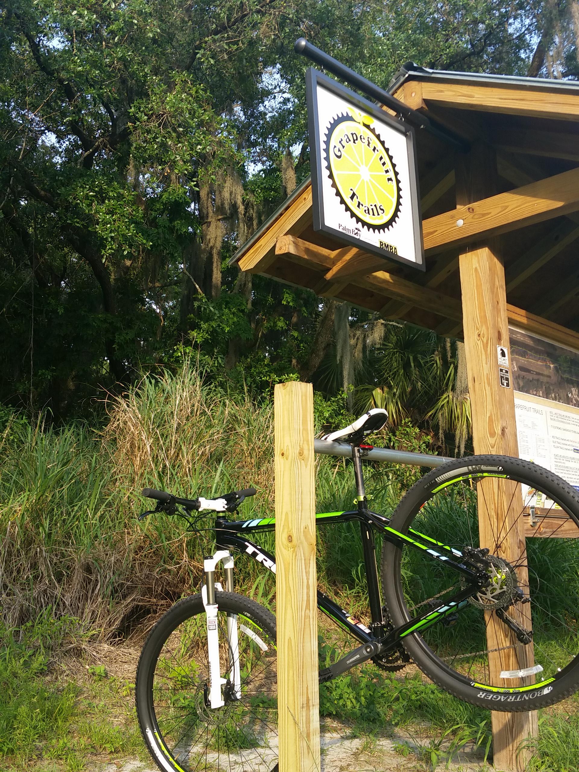 Trek X-Caliber 7: A mountain bike leaning against a wooden post at the entrance of Grapefruit Trails, with a sign hanging above that features a yellow gear and the trail name. Lush greenery and trees are in the background, suggesting a natural outdoor setting.