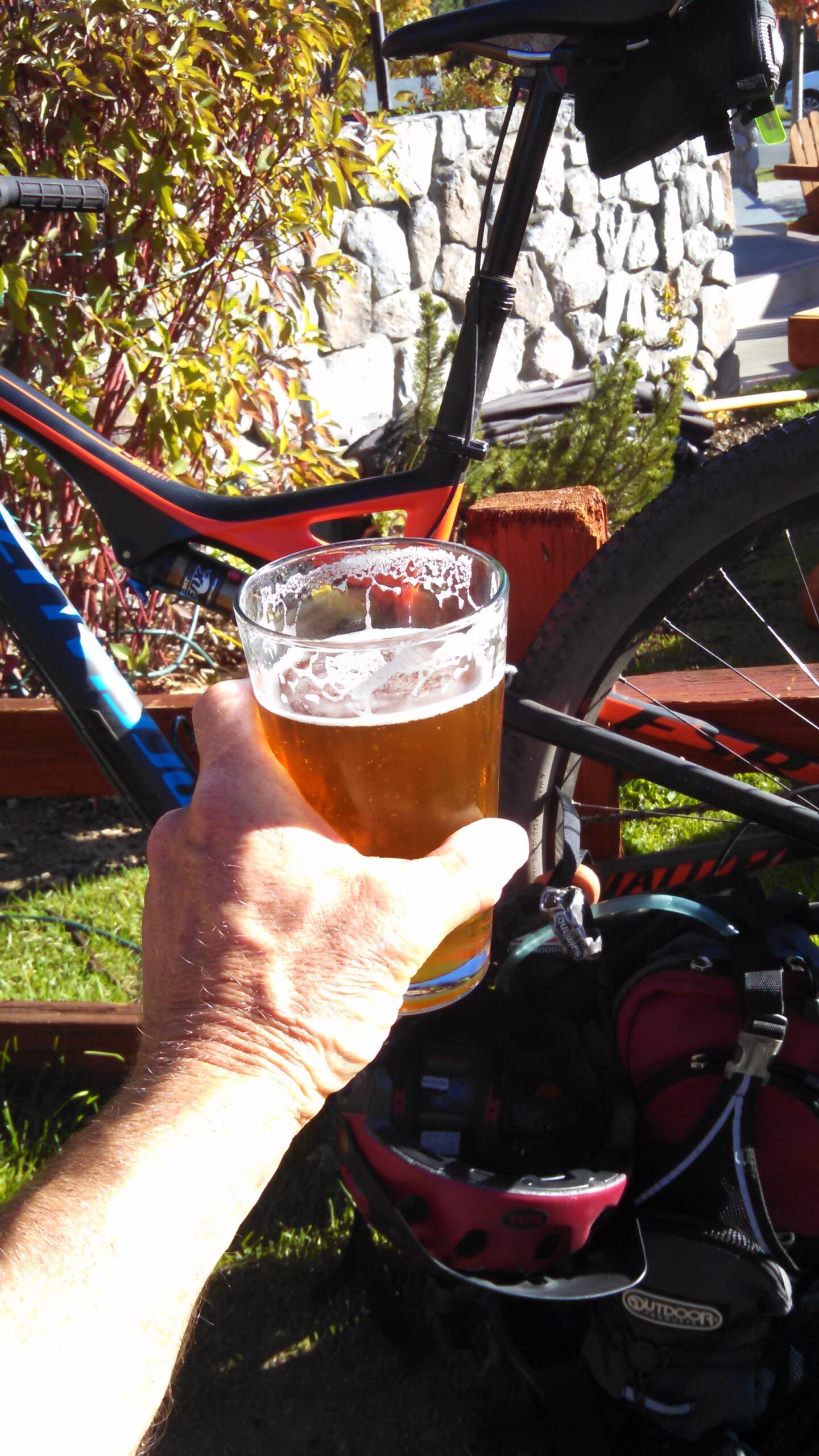 Specialized Stumpjumper FSR Expert Carbon EVO 29: A close-up of a hand holding a glass of beer, with a mountain bike and a backpack in the background. The scene is set outdoors, showcasing greenery and a stone wall. The beer has a frothy head and a golden color, suggesting a refreshing post-ride drink.