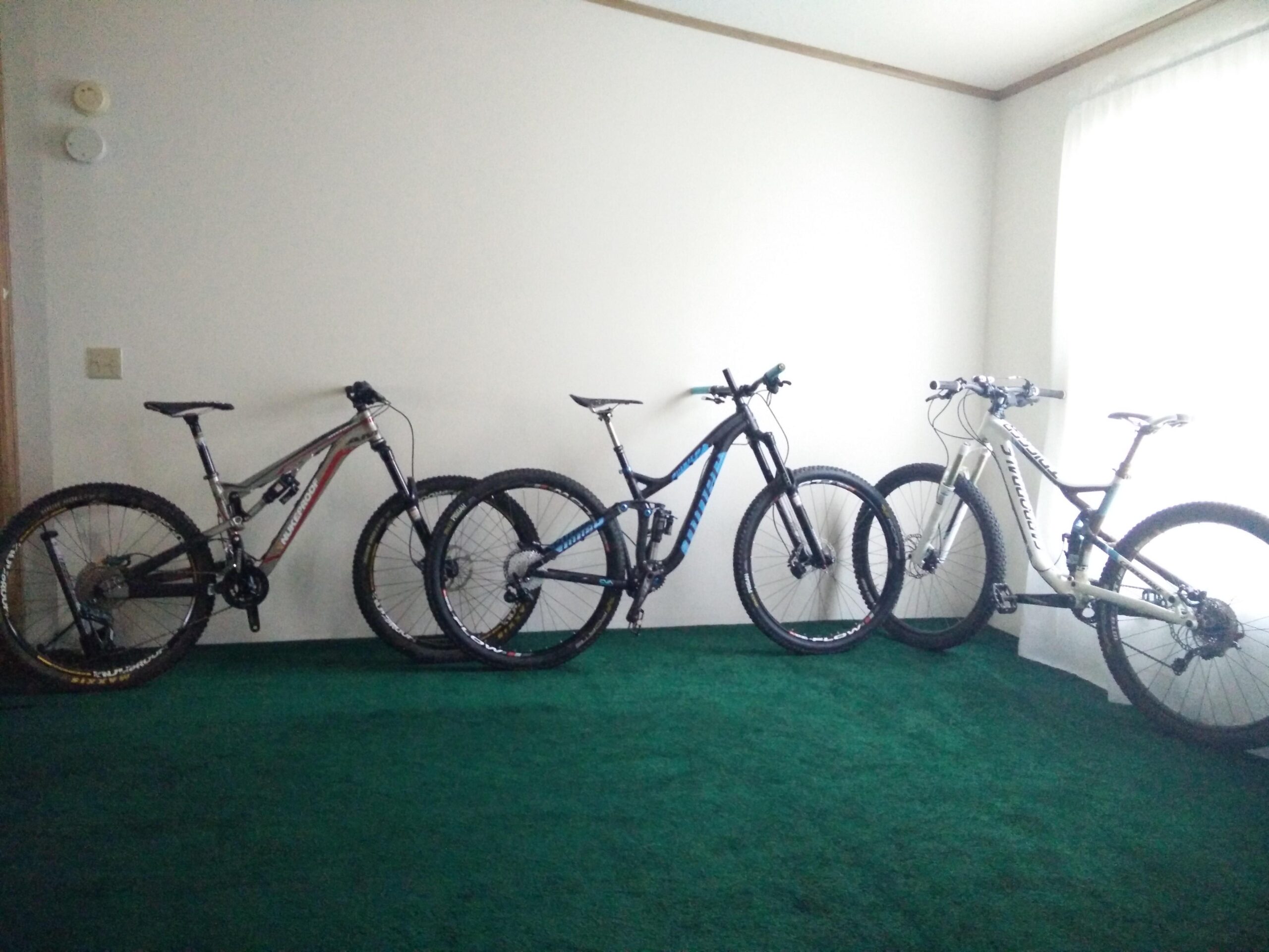 Nukeproof Mega AM 275 Comp: Three mountain bikes are lined up against a wall in a room with green carpet. The bikes vary in color and design, showcasing a mix of styles suitable for off-road riding. A large window with white curtains lets in natural light, illuminating the scene.