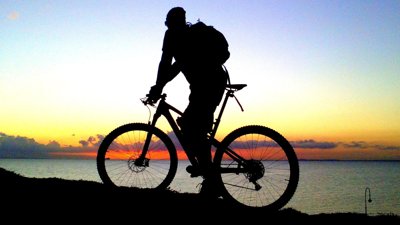 Silhouette of a cyclist standing beside a mountain bike, overlooking a sunset over the ocean, with a gradient sky in twilight hues. Yarra Trails mountain bike trail.