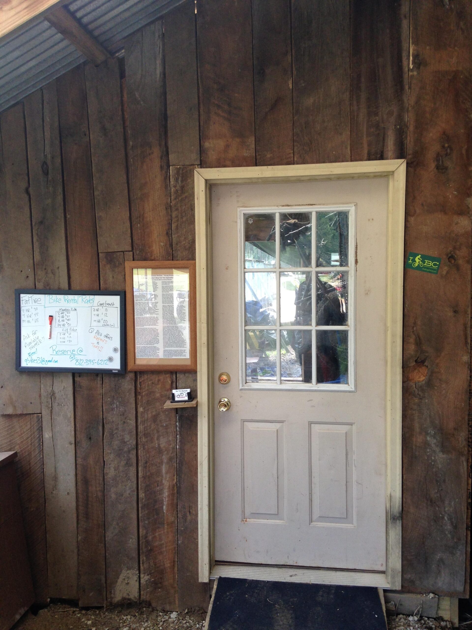 A rustic wooden building featuring a white door with a glass panel, a dark mat at the entrance, and a small signboard with information related to bike rentals. The exterior walls are made of weathered wooden planks, giving it an earthy appearance. A framed document is visible next to the door, alongside a chalkboard listing rental details.